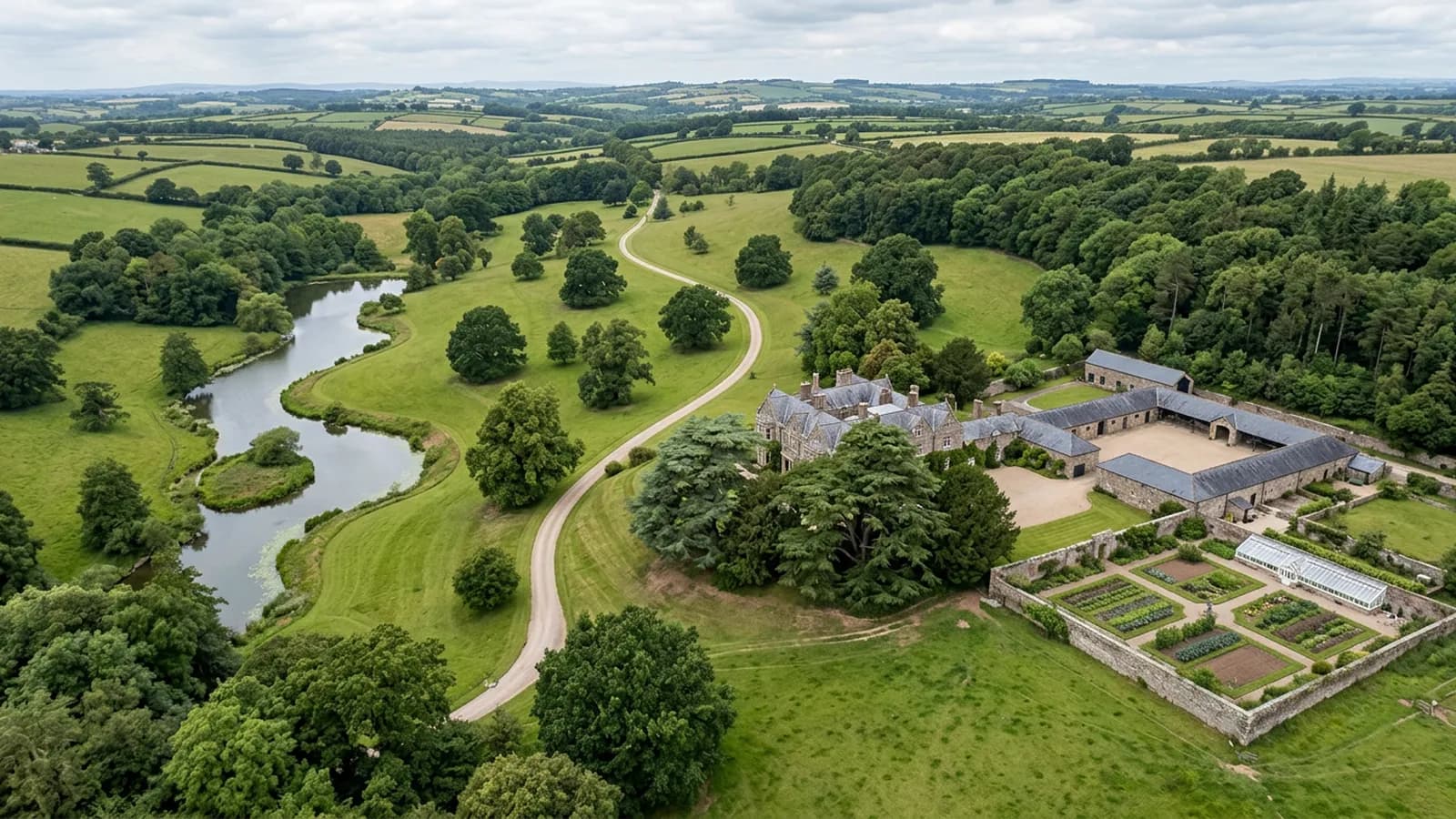 A country estate in Devon from the air, parkland grass with mature standing trees, a serpentine lake, a long drive and stable ranges under slate