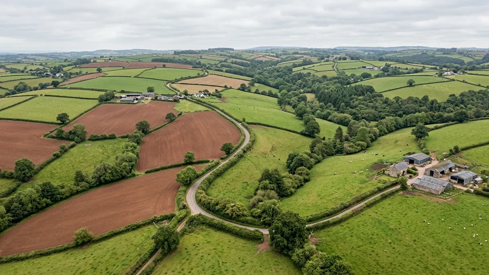 Mid Devon farmland from the air, a patchwork of hedged fields over rolling hills with red soil ploughland beside grass leys and scattered farmsteads