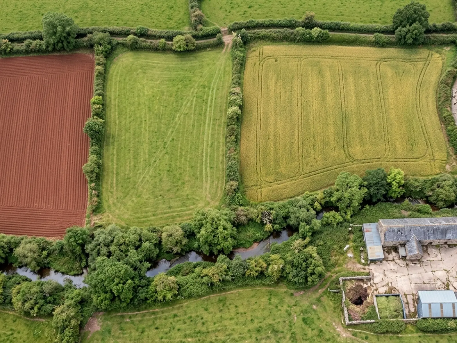 Hedged Devon field parcels photographed straight down from height, one freshly ploughed to red soil, one in grass and one under crop, with a farm track and a stone barn in the corner